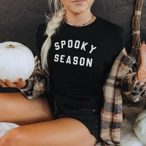 Puede incluir: Una mujer con una camiseta negra con el texto "Spooky Season" en letras blancas. Est&aacute; sosteniendo una calabaza blanca y lleva una camisa de cuadros.