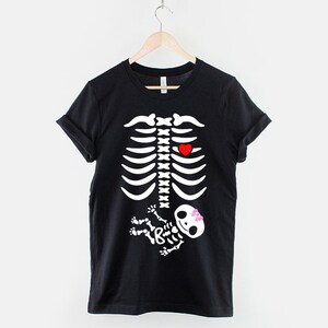 May include: Black t-shirt with a white skeleton graphic. The skeleton has a red heart in its chest and a small white skeleton baby in its belly with a pink bow.