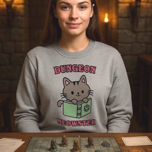 Può includere: Una persona indossa una felpa grigia con la scritta "DUNGEON MEOWSTER" e un gatto dei cartoni animati che legge un libro. Un gioco di ruolo da tavolo è allestito di fronte a lei, con dadi, miniature e carte di gioco.