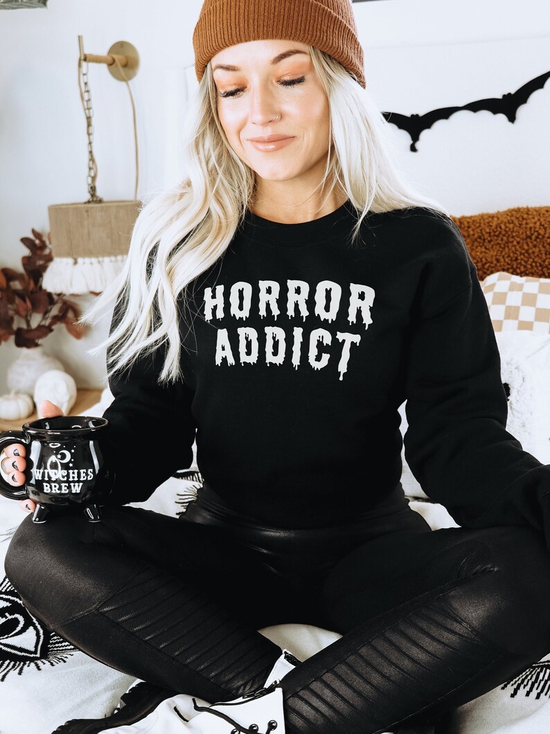 Puede incluir: Una mujer lleva una sudadera negra con el texto "HORROR ADDICT" en letras blancas que gotean. Est&aacute; sentada en una cama con una taza negra que dice "WITCHES BREW" en letras blancas.