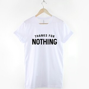 May include: White t-shirt with the phrase "THANKS FOR NOTHING" printed in bold black letters. The shirt is hanging on a wooden hanger suspended from a metal chain. The t-shirt has short sleeves and a crew neck.