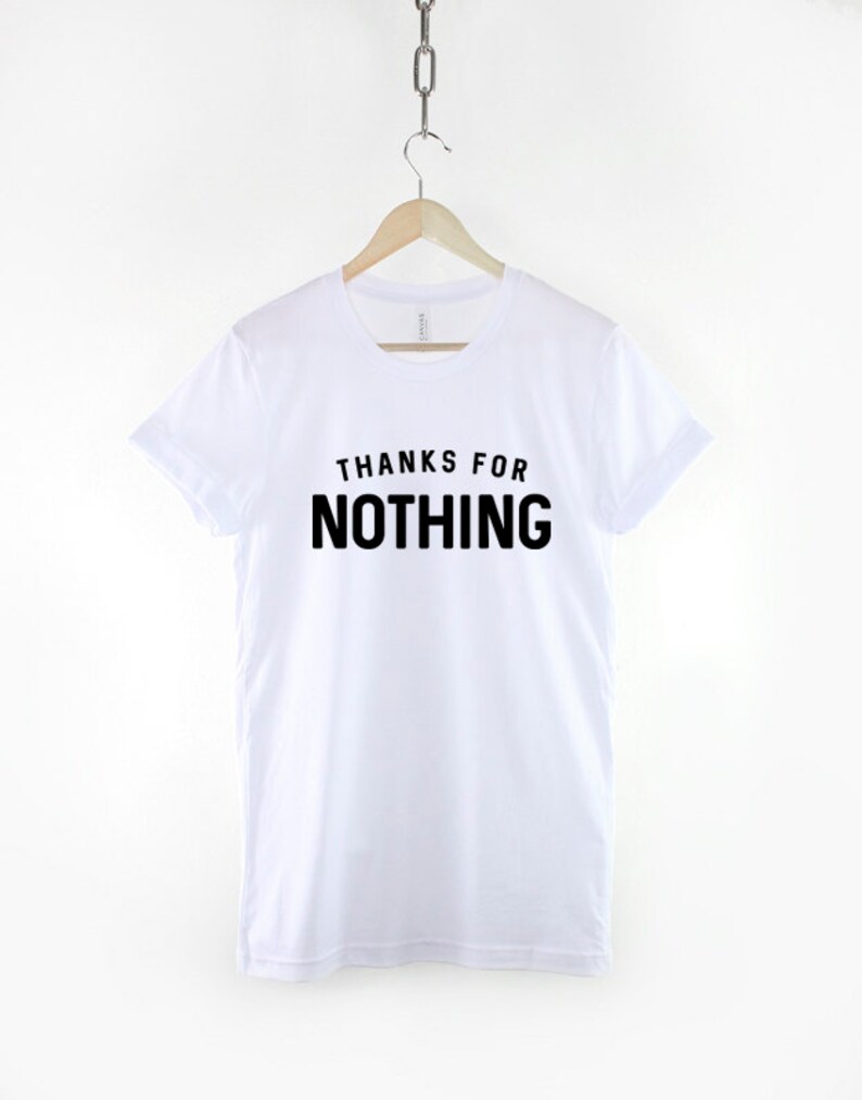May include: White t-shirt with the phrase "THANKS FOR NOTHING" printed in bold black letters. The shirt is hanging on a wooden hanger suspended from a metal chain. The t-shirt has short sleeves and a crew neck.