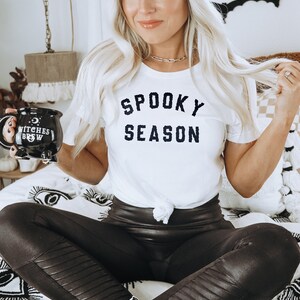 Puede incluir: Una mujer con una camiseta blanca con el texto "Spooky Season" en letras negras, leggings de cuero negro y un gorro marr&oacute;n. Est&aacute; sosteniendo una taza negra con el texto "Witches Brew" en letras blancas.