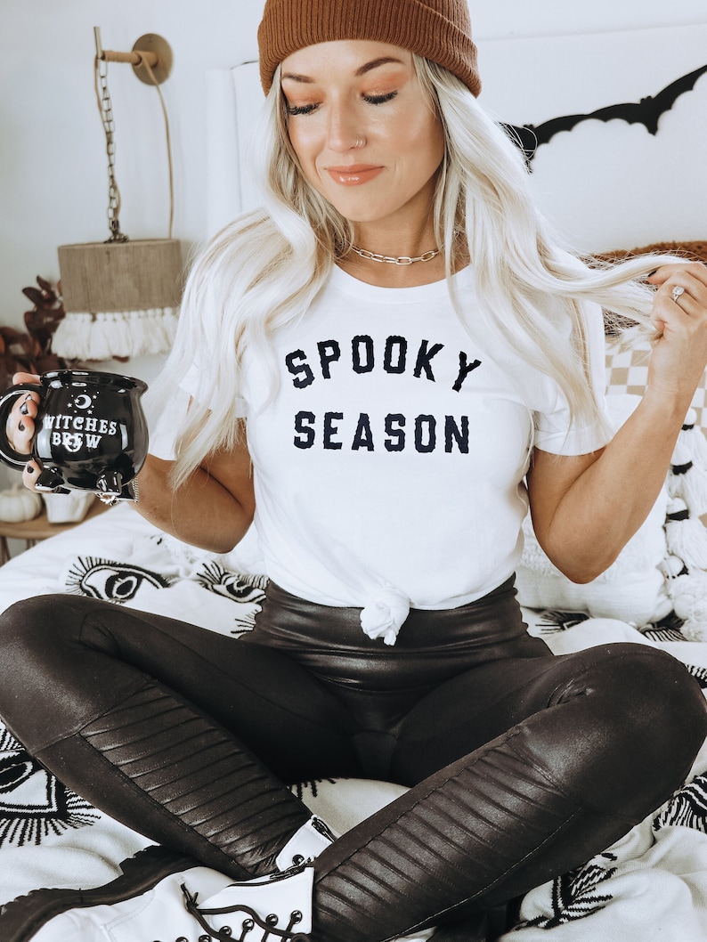 Puede incluir: Una mujer con una camiseta blanca con el texto "Spooky Season" en letras negras, leggings de cuero negro y un gorro marr&oacute;n. Est&aacute; sosteniendo una taza negra con el texto "Witches Brew" en letras blancas.
