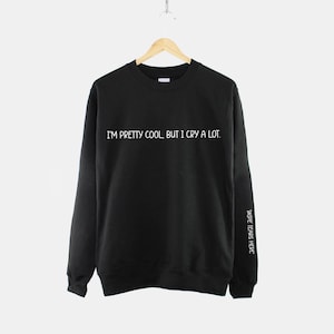 May include: Black sweatshirt with the text "I'm pretty cool, but I cry a lot." printed in white on the front. The text "Wide tears here" is printed in white on the right sleeve.