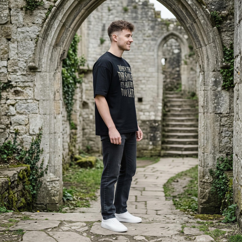 May include: A black short-sleeved t-shirt with white text that reads "WHEN THE DAY SMILES IT'S ALREADY TOO LATE" paired with dark gray jeans and white sneakers. The t-shirt features a crew neckline and a relaxed fit, worn with slim-fit denim pants.