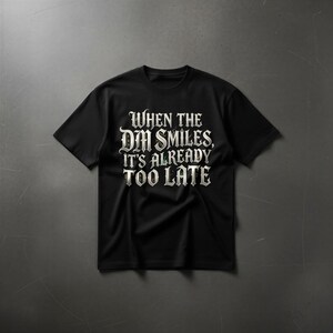 May include: A black short-sleeved t-shirt featuring white gothic-style lettering on the chest that reads, "WHEN THE DM SMILES, IT'S ALREADY TOO LATE." The text is centered and printed in a bold, decorative font with a slight weathered texture.