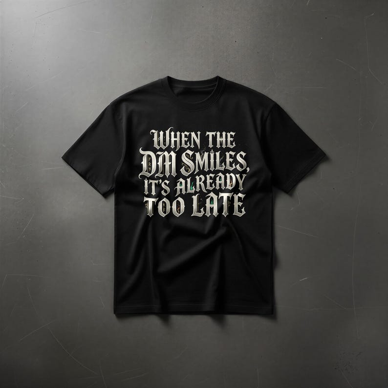 May include: A black short-sleeved t-shirt featuring white gothic-style lettering on the chest that reads, "WHEN THE DM SMILES, IT'S ALREADY TOO LATE." The text is centered and printed in a bold, decorative font with a slight weathered texture.