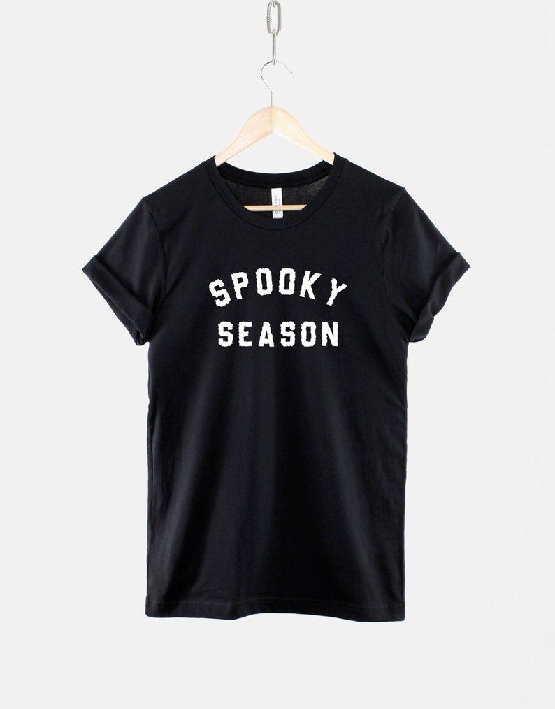 Puede incluir: Camiseta negra con texto blanco que dice "Spooky Season" en una fuente universitaria vintage.