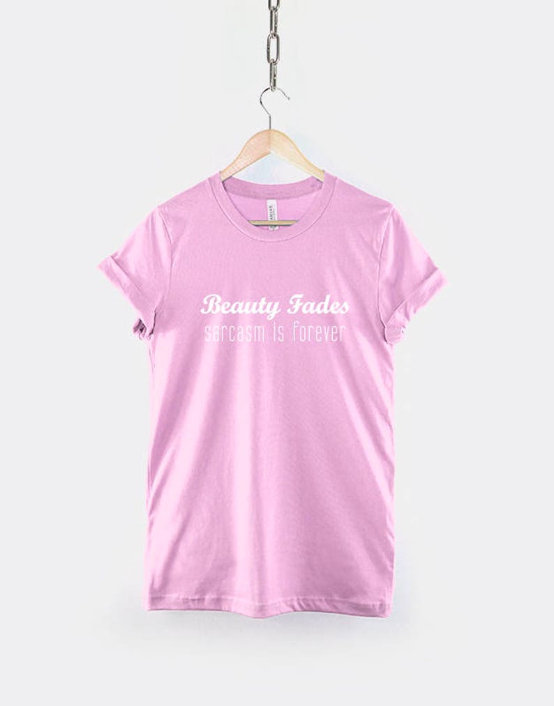 May include: Pink t-shirt with the text "Beauty Fades sarcasm is forever" in white.