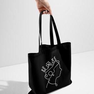 Op de afbeelding: Zwarte tote bag met een witte lijntekening van een vrouwenhoofd met een kroon.