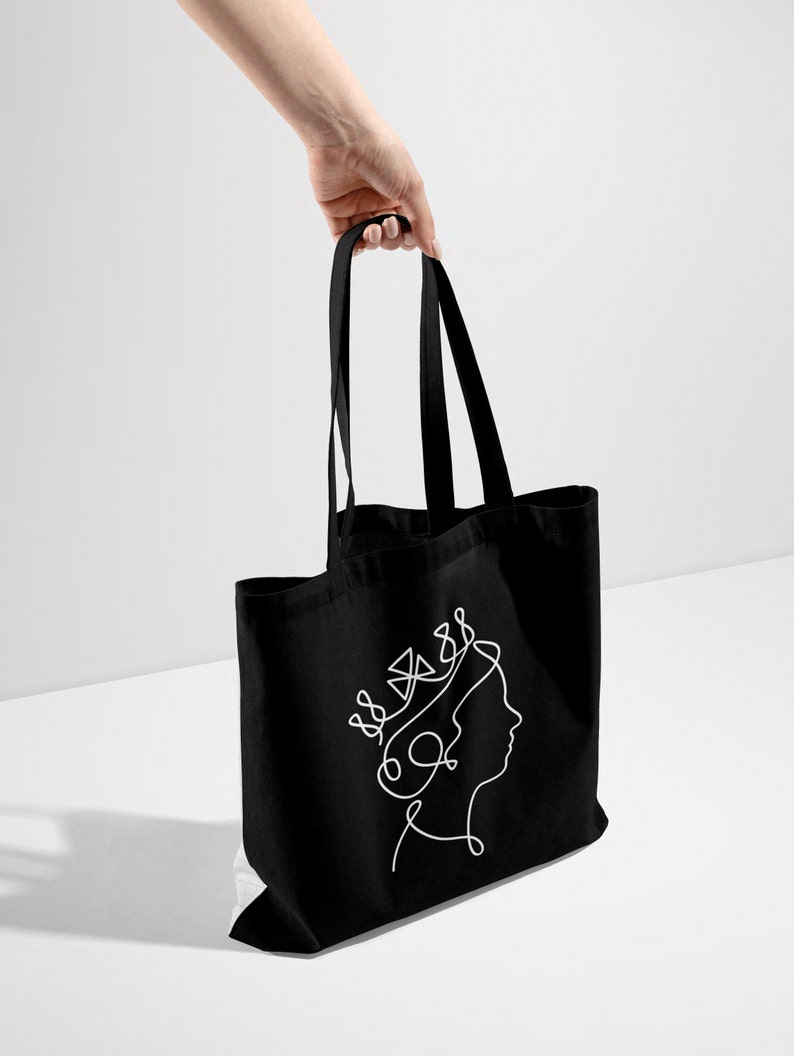 Op de afbeelding: Zwarte tote bag met een witte lijntekening van een vrouwenhoofd met een kroon.