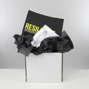 May include: A white ceramic mug with a black handle is packaged in a white box with black tissue paper. The box has a black card on top with the text "RESILIENT" and a website address.