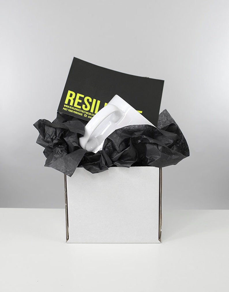 May include: A white ceramic mug with a black handle is packaged in a white box with black tissue paper. The box has a black card on top with the text "RESILIENT" and a website address.