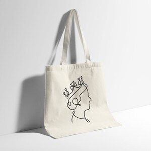 Op de afbeelding: Een witte canvas tote bag met een zwarte lijntekening van een vrouwenhoofd met een kroon op haar hoofd.