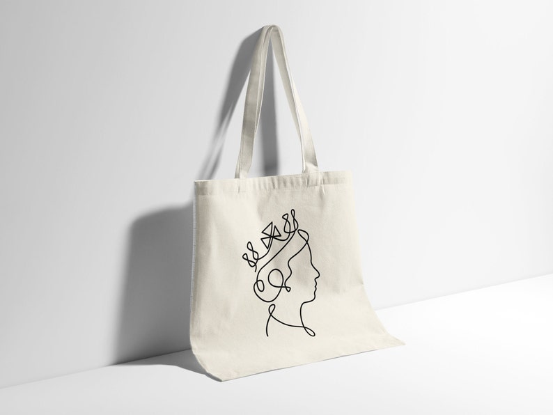 Op de afbeelding: Een witte canvas tote bag met een zwarte lijntekening van een vrouwenhoofd met een kroon op haar hoofd.