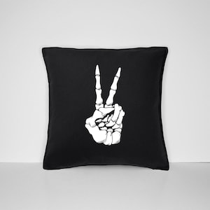 May include: Black throw pillow with a white skeleton hand making the peace sign.