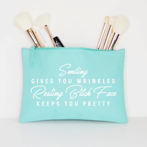 May include: A light teal makeup bag with a zipper, filled with makeup brushes. The bag has the text "Smiling gives you wrinkles, Resting Bitch Face keeps you pretty" in white script.