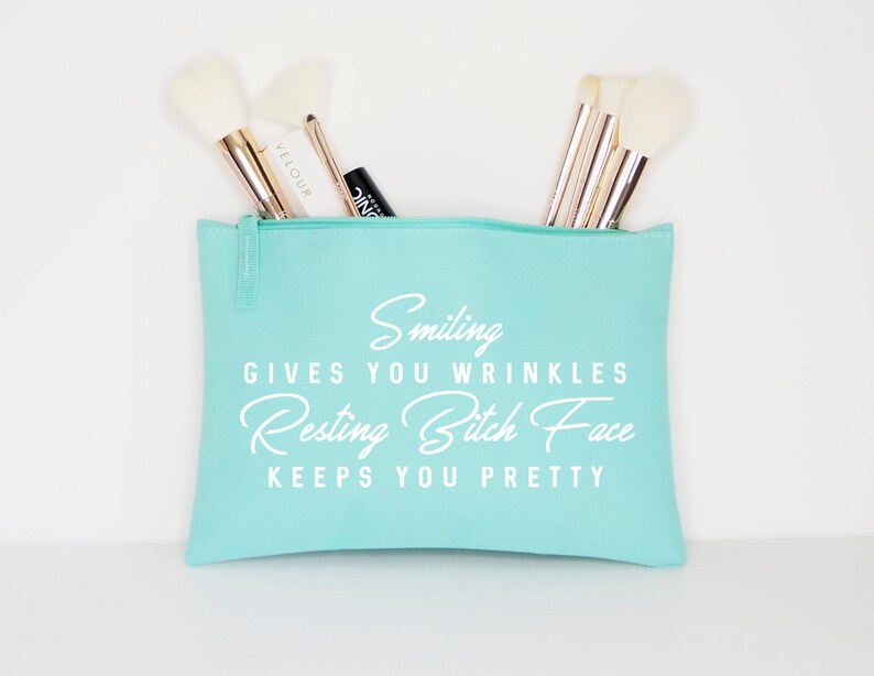 May include: A light teal makeup bag with a zipper, filled with makeup brushes. The bag has the text "Smiling gives you wrinkles, Resting Bitch Face keeps you pretty" in white script.