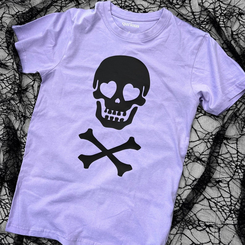 May include: A lavender t-shirt with a black skull and crossbones design. The skull has heart-shaped eyes.