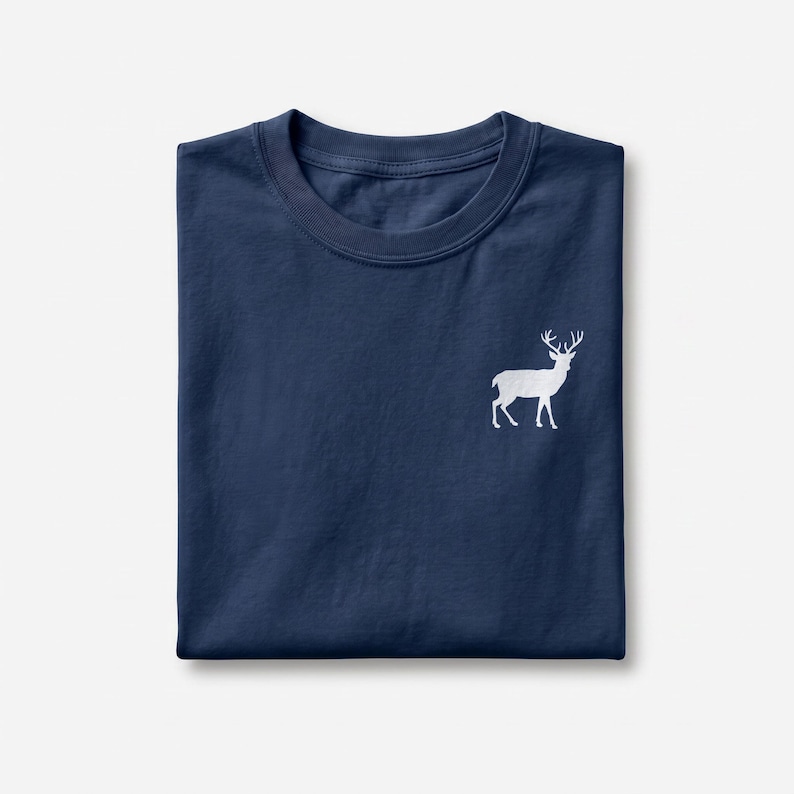 Puede incluir: Una camiseta azul marino de cuello redondo con un peque&ntilde;o gr&aacute;fico blanco minimalista de un ciervo de pie con astas, situado en la parte superior derecha del pecho. La prenda est&aacute; doblada cuidadosamente, mostrando el cuello y la textura.