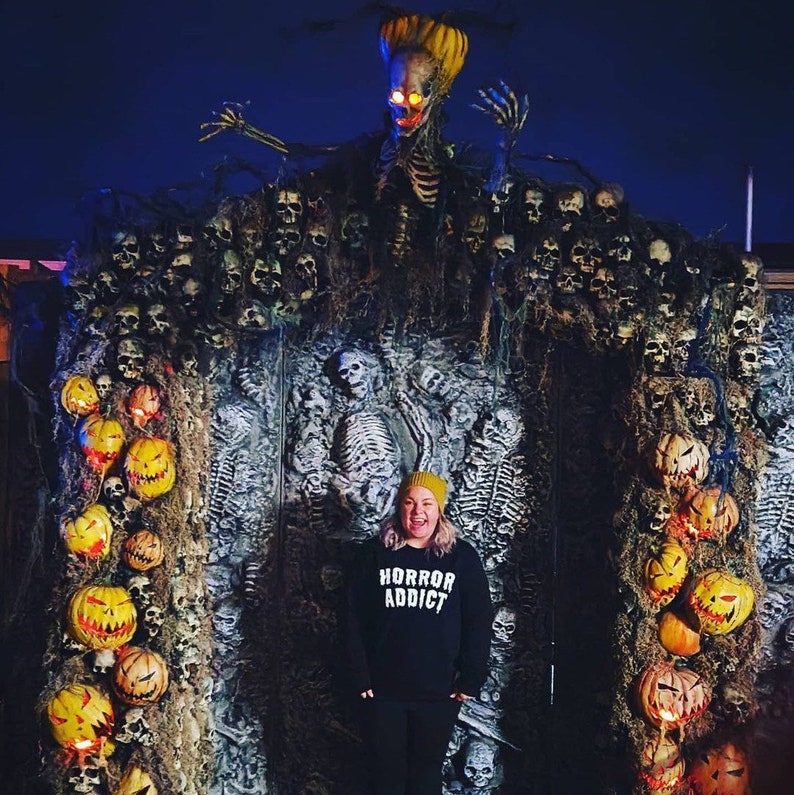 Puede incluir: Una persona con una sudadera negra con el texto "Horror Addict" en letras blancas, un gorro amarillo, y de pie frente a un tel&oacute;n de fondo de tem&aacute;tica de Halloween. El tel&oacute;n de fondo presenta un esqueleto con ojos brillantes, una pared de cr&aacute;neos y una guirnalda de calabazas.