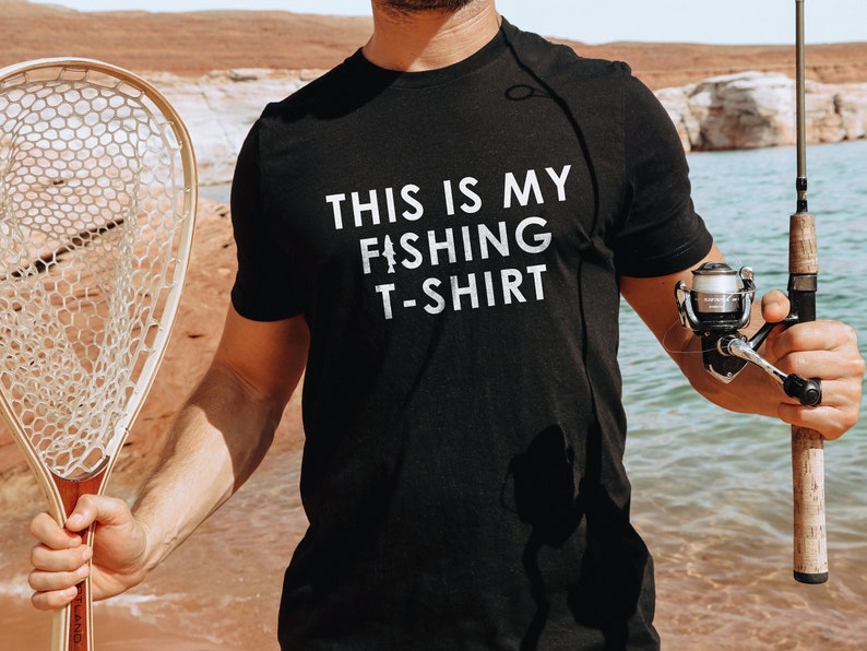 Może przedstawiać: Czarny t-shirt z białym napisem "This is my fishing t-shirt". Koszulkę nosi osoba trzymająca wędkę i siatkę.