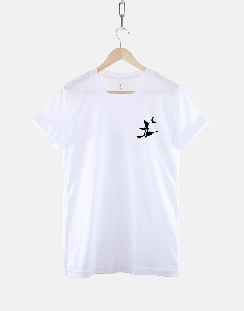 Op de afbeelding: Wit T-shirt met een zwart silhouet van een heks die op een bezem vliegt met een halve maan op de achtergrond.