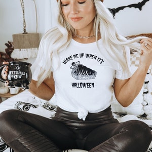 Puede incluir: Una mujer con una camiseta blanca con el texto "Wake me up when it's Halloween" y una ilustraci&oacute;n en blanco y negro de un esqueleto en un ata&uacute;d. Tambi&eacute;n lleva pantalones de cuero negro y una boina marr&oacute;n.