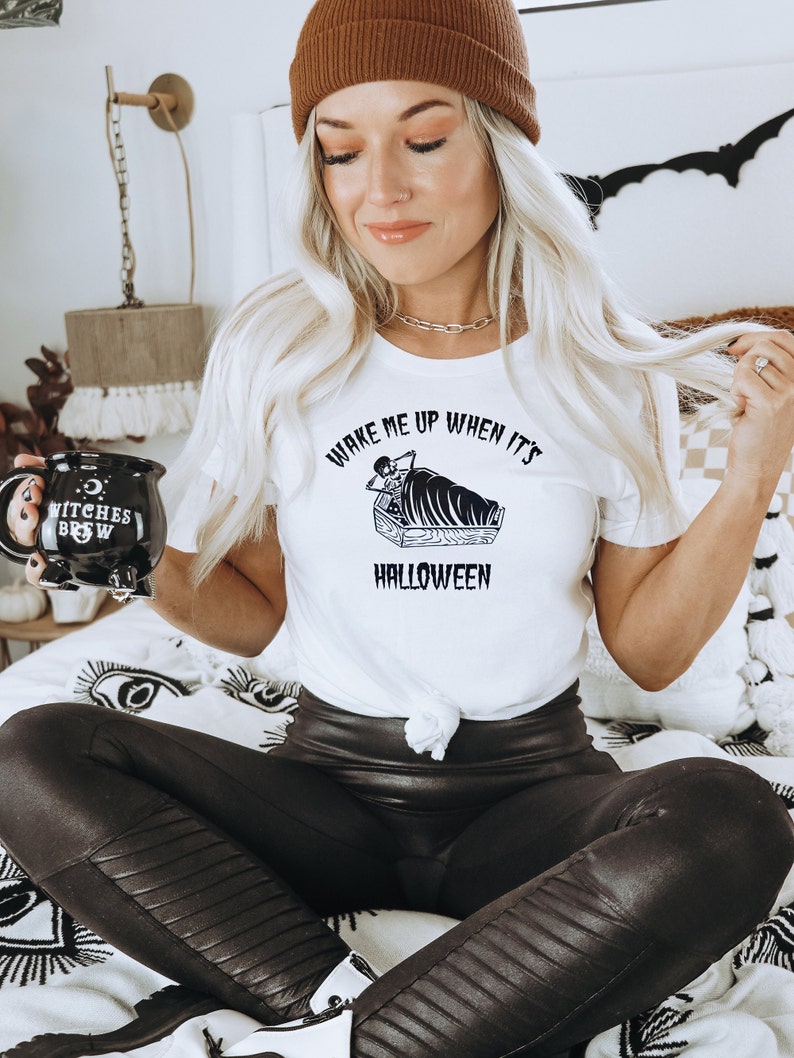 Puede incluir: Una mujer con una camiseta blanca con el texto "Wake me up when it's Halloween" y una ilustraci&oacute;n en blanco y negro de un esqueleto en un ata&uacute;d. Tambi&eacute;n lleva pantalones de cuero negro y una boina marr&oacute;n.