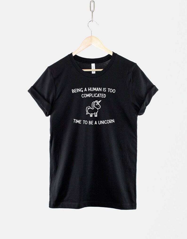 May include: Black t-shirt with white text that reads "Being a human is too complicated. Time to be a unicorn." A small unicorn illustration is above the text.