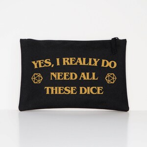 Puede incluir: Bolsa negra con cremallera y texto dorado: "YES, I REALLY DO NEED ALL THESE DICE". Dos formas geom&eacute;tricas doradas flanquean el texto. La bolsa es rectangular y parece estar hecha de lona. El fondo es blanco.