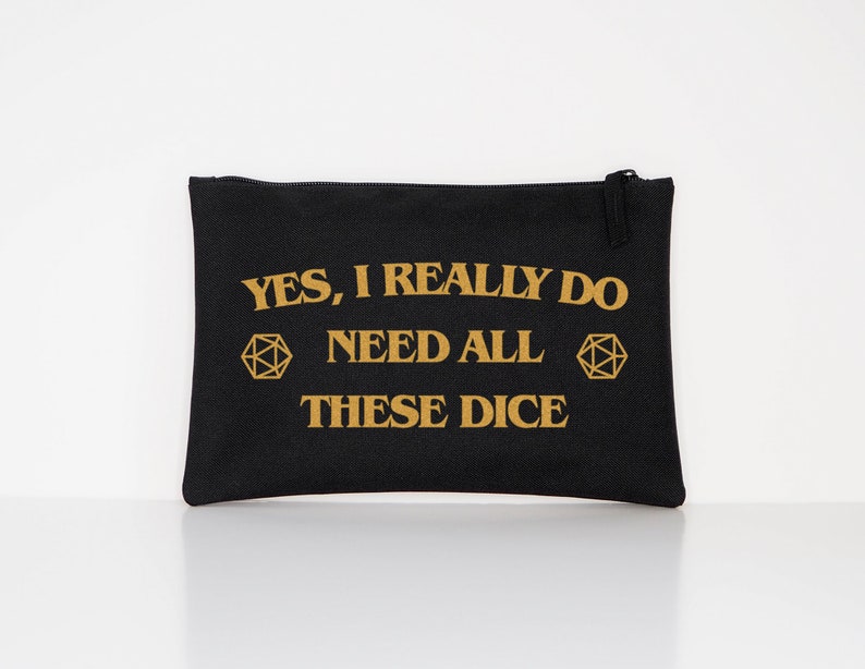 Puede incluir: Bolsa negra con cremallera y texto dorado: "YES, I REALLY DO NEED ALL THESE DICE". Dos formas geom&eacute;tricas doradas flanquean el texto. La bolsa es rectangular y parece estar hecha de lona. El fondo es blanco.