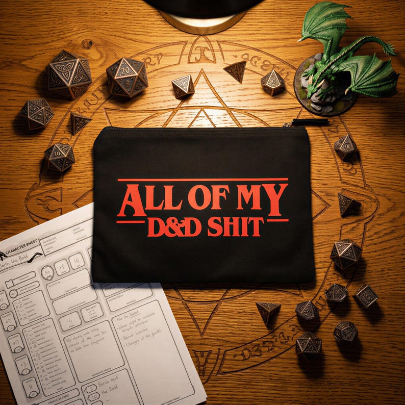 May include: A black rectangular zippered pouch featuring the red text ALL OF MY D&D SHIT centred on the front. The pouch is surrounded by various metallic polyhedral dice, a white paper character sheet, and a small green dragon figurine.
