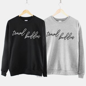 K&ouml;nnte beinhalten: Zwei Sweatshirts, eines schwarz und eines grau, mit dem Text "travel buddies" in wei&szlig;er Schrift auf der Vorderseite.