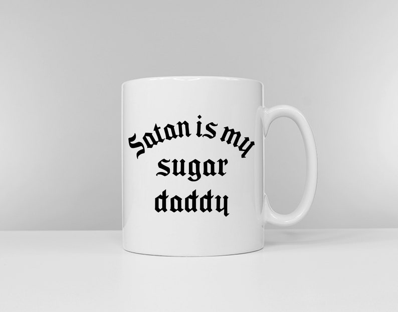 Puede incluir: Taza de cer&aacute;mica blanca con un dise&ntilde;o de fuente g&oacute;tica negra que dice "Satan is my sugar daddy".