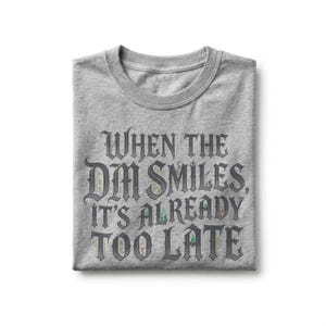 May include: A heather gray crew neck t-shirt featuring dark gray gothic-style lettering that reads, "WHEN THE DM SMILES, IT'S ALREADY TOO LATE." The text includes small, colorful, intricate illustrations of fantasy-themed items within the letters.