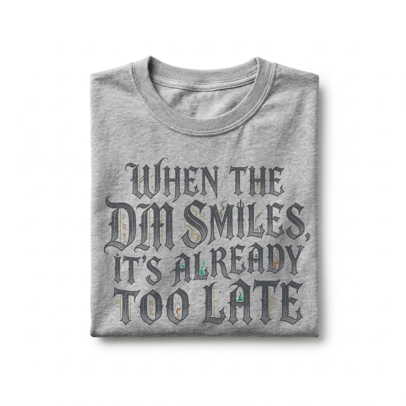 May include: A heather gray crew neck t-shirt featuring dark gray gothic-style lettering that reads, "WHEN THE DM SMILES, IT'S ALREADY TOO LATE." The text includes small, colorful, intricate illustrations of fantasy-themed items within the letters.