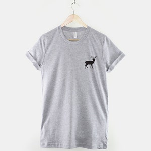 Puede incluir: Una camiseta gris jaspeado de manga corta con una peque&ntilde;a silueta negra de un ciervo con astas en el lado izquierdo del pecho. La prenda tiene cuello redondo y mangas remangadas, colgada de una percha de madera sobre un fondo liso.
