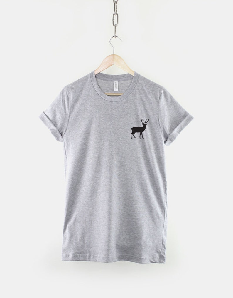 Puede incluir: Una camiseta gris jaspeado de manga corta con una peque&ntilde;a silueta negra de un ciervo con astas en el lado izquierdo del pecho. La prenda tiene cuello redondo y mangas remangadas, colgada de una percha de madera sobre un fondo liso.