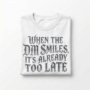 May include: A white short-sleeved t-shirt featuring the dark grey text "WHEN THE DM SMILES, IT'S ALREADY TOO LATE" in a stylized gothic font. The lettering includes small, subtle decorative elements and flourishes integrated into the characters.