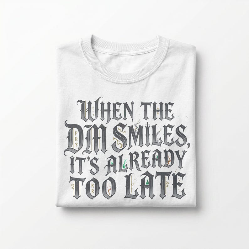 May include: A white short-sleeved t-shirt featuring the dark grey text "WHEN THE DM SMILES, IT'S ALREADY TOO LATE" in a stylized gothic font. The lettering includes small, subtle decorative elements and flourishes integrated into the characters.