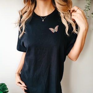 May include: A black t-shirt featuring a colourful butterfly graphic on the front. The butterfly has pink, yellow and blue wings.