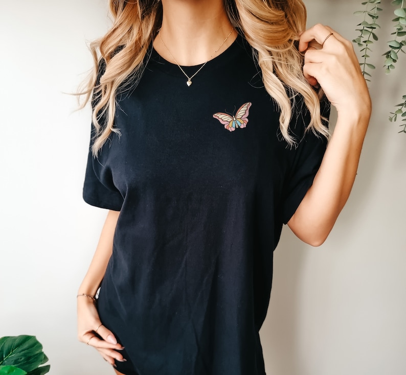 May include: A black t-shirt featuring a colourful butterfly graphic on the front. The butterfly has pink, yellow and blue wings.