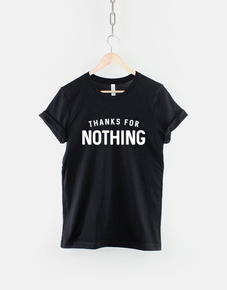 May include: Black t-shirt with the phrase "THANKS FOR NOTHING" printed in white, displayed on a wooden hanger. The shirt is hanging from a metal chain against a white background. The sleeves are cuffed.