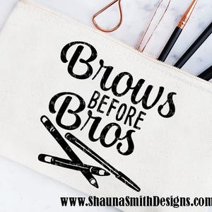 May include: A white canvas makeup bag with a zipper closure. The bag has the text "Brows Before Bros" printed in black. The bag is filled with makeup brushes and a lash curler.