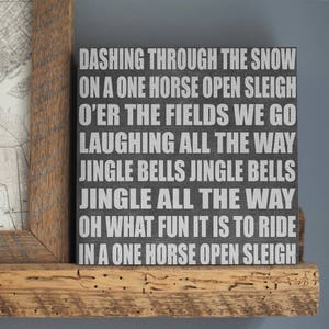 May include: A black wooden sign with white lettering, featuring the lyrics "Dashing through the snow...Jingle Bells...Oh what fun it is to ride in a one horse open sleigh." The sign is displayed on a wooden shelf.