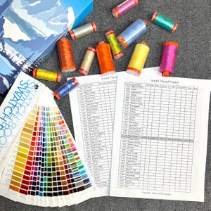 Könnte beinhalten: Eine Nahaufnahme eines Faden-Tracker-Diagramms mit einem Farbmusterbuch und Spulen mit Stickgarn in verschiedenen Farben. Das Diagramm ist mit "Aurifil Thread Tracker" beschriftet und enthält Spalten für die Fadennummer, den Farbnamen und die verwendete Fadenmenge. Das Farbmusterbuch ist mit "Swatch Book" beschriftet und zeigt eine Vielzahl von Farben.