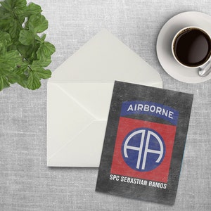 May include: A black card with a red, white, and blue military insignia. The insignia is a circle with the letters "AA" inside. The text "AIRBORNE" is above the insignia and the text "SPC SEBASTIAN RAMOS" is below the insignia.