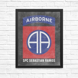 May include: A black framed print with a red, white, and blue military insignia. The insignia is a circle with the letters "AA" in white inside. The text "AIRBORNE" is above the circle and "SPC SEBASTIAN RAMOS" is below the circle.